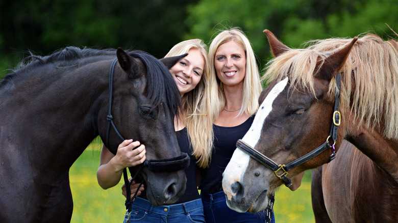 A mature blonde woman and blonde Gen Z woman smiling while standing between two horses in a pasture