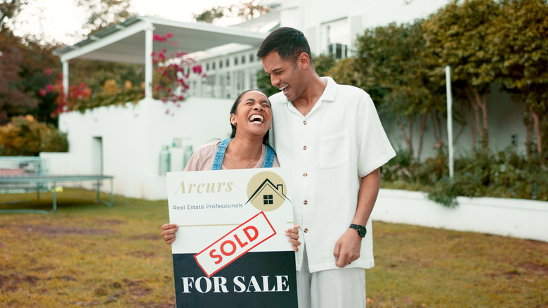 A smiling, happy couple in front of a home with sold sign on the front lawn