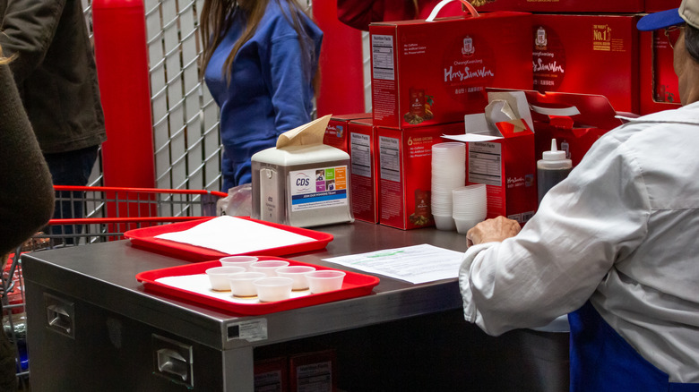 An elderly Costco sampler preps cups of free drinks at a busy store.