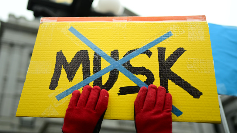 Gloved hands holding a handmade anti-Elon Musk sign.