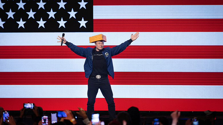 Elon Musk on stage in front of a U.S. flag, wearing a cheesehead hat.