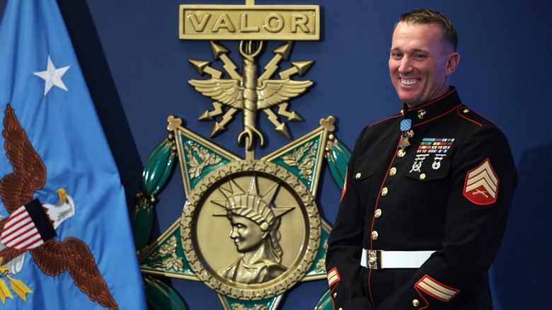A U.S. Marine in dress uniform smiles while wearing the Medal of Honor, standing beside a large Medal of Honor emblem and a U.S. flag