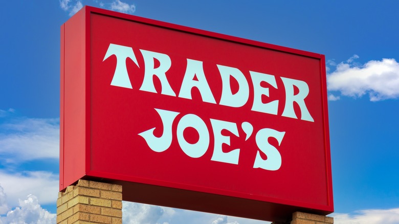 A cheery red Trader Joe's sign on brick posts, in front of a bright blue sky dotted with puffy white clouds.
