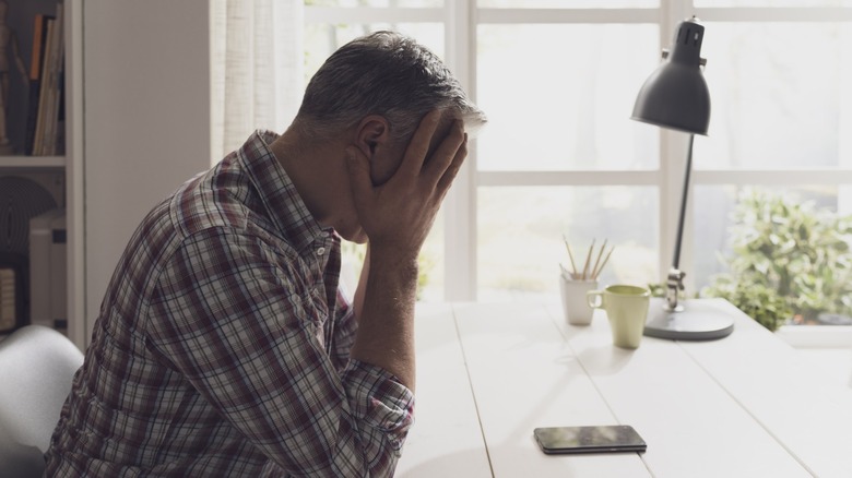 A man in a plaid shirt holding his head in his hands sitting at a table in front of a cell phone.