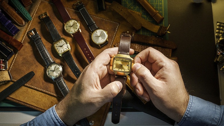 A pair of hands holds a watch with a brown leather band and a rectangular gold-colored face with various other watches of varying colors and styles in the background.