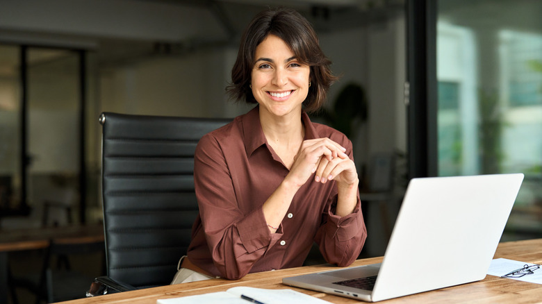 A smiling woman sitting at a desk with an open laptop in front of her.