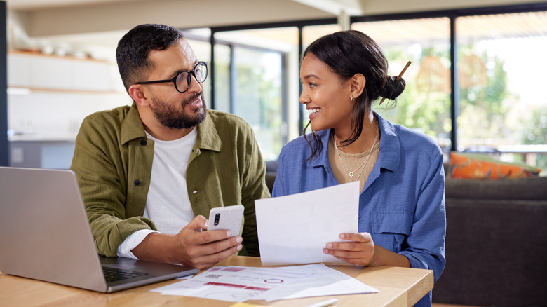 A happy couple sitting at a table looking at each other while the man is holding a phone and the woman has a paper in her hand with an open laptop to one side.