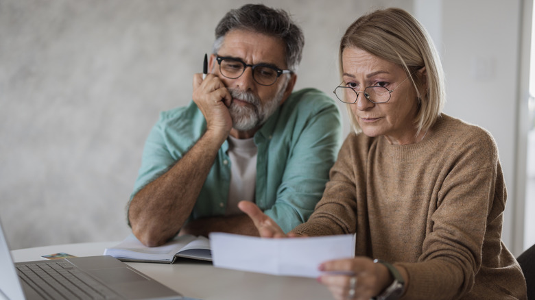 A retired couple sitting at a table looking at a paper with worried expressions and an open laptop in the background.