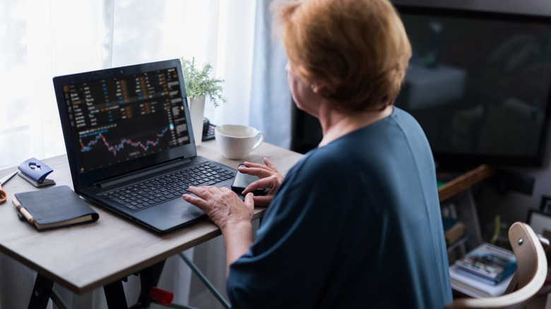 A mature woman sitting at a desk while monitoring her investments on a laptop