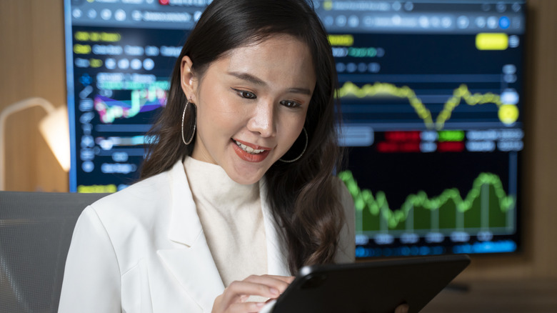 A smiling Millennial woman in front of a stock monitor reading a tablet.