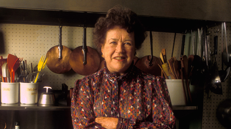 Julia Child standing with her arms crossed in a kitchen with lots of cooking utensils behind her.