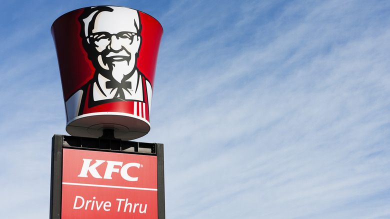 A large KFC bucket bearing the image of Colonel Sanders sits atop a drive-thru sign, with a bright blue sky in the background.