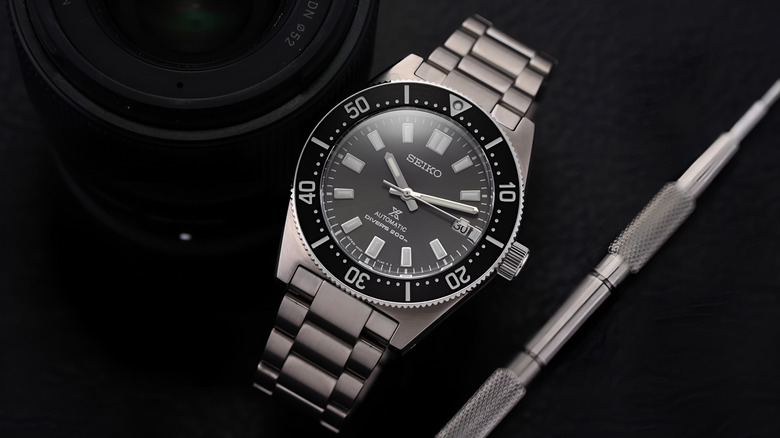 The Seiko SPB143 on a flat black surface and at its side, what looks like a camera lens and a pen