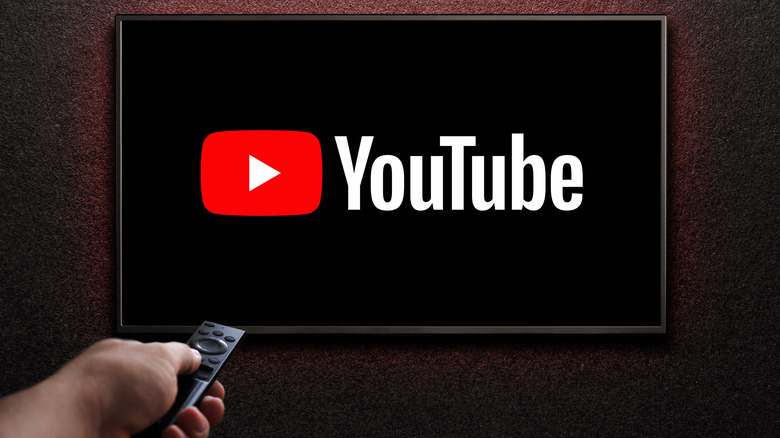 A TV with the red YouTube logo and white words on a black screen and a hand holding a remote pointing at the TV in the bottom left corner.