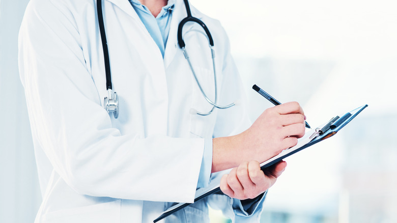 A medium shot of a doctor's midsection, wearing white coat and stethoscope, hands holding a pen hovering over a clipboard, against a bright white clinical background.