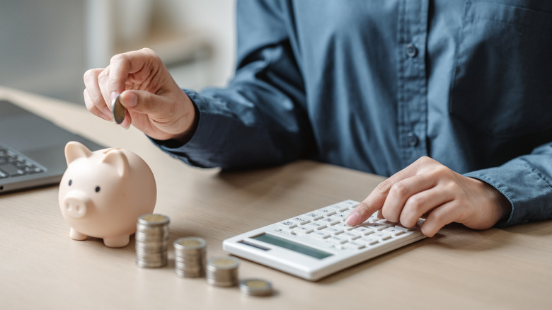 Close up of seated male torso where wan is putting coin in piggy bank and typing on calculator