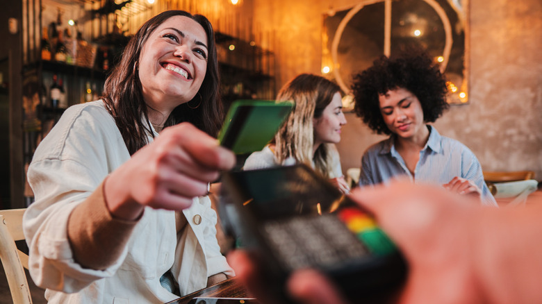 Smiling woman sitting at bar pays with credit card while two women sit in background talking