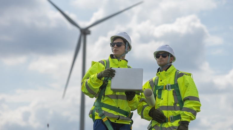 Two wind turbine technicians wearing protective gear standing with a laptop outside in front of a wind turbine.