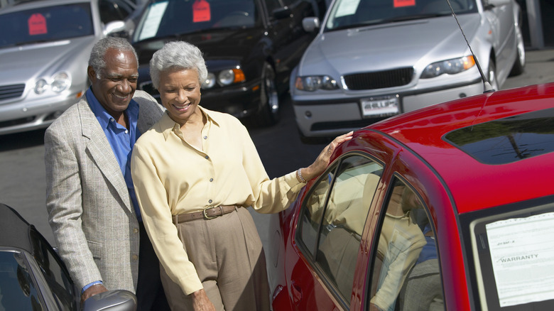 A smiling retired couple at a car lot looking at a red car with other vehicles in the background.
