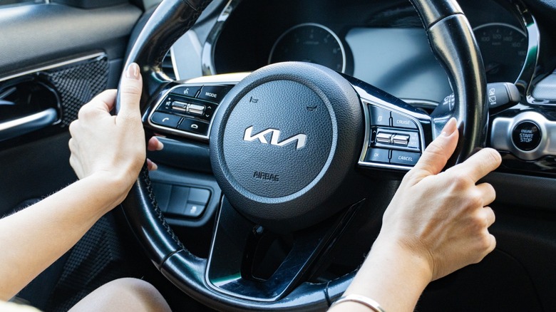 A driver grips a Kia steering wheel, with dashboard controls and the push-button ignition visible