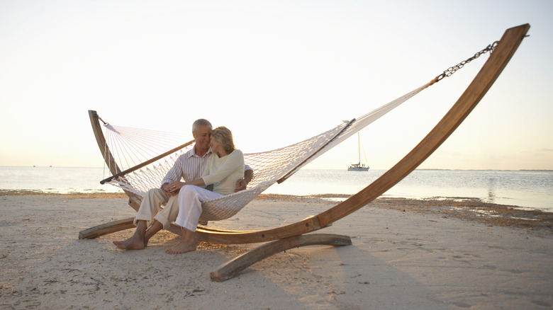 A retired couple sitting in a hammock on the beach with a sailboat in the background.