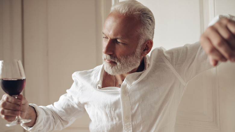 A fashionable mature man looking at a glass of wine in his hand whil leaning against a wall.