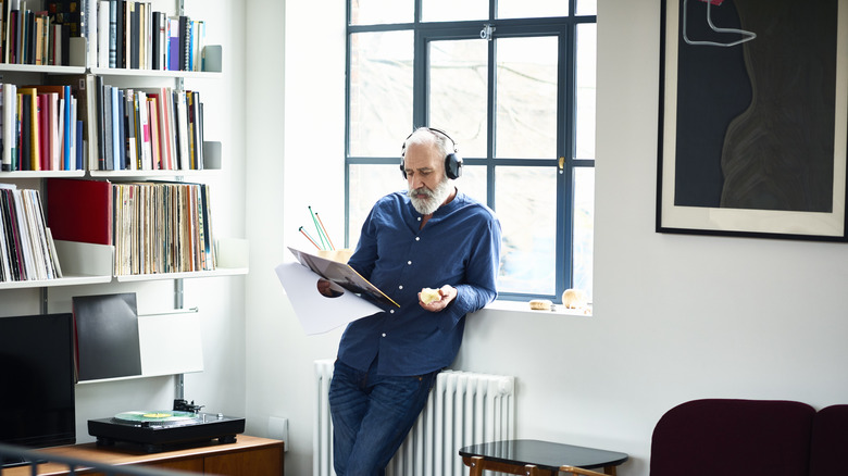 A mature man in his home wearing headphones while reading a record sleeve.
