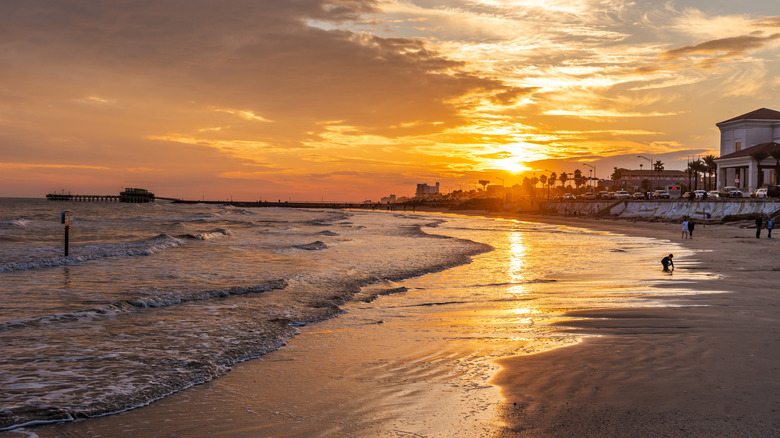 A beach scene at sunset with a pier, buildings, and people in the background.