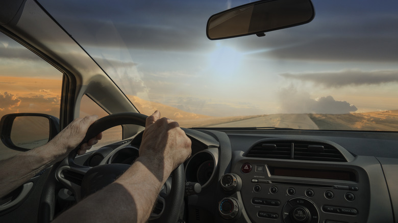 A man's hands at the wheel of a car in a road in the desert at sunset