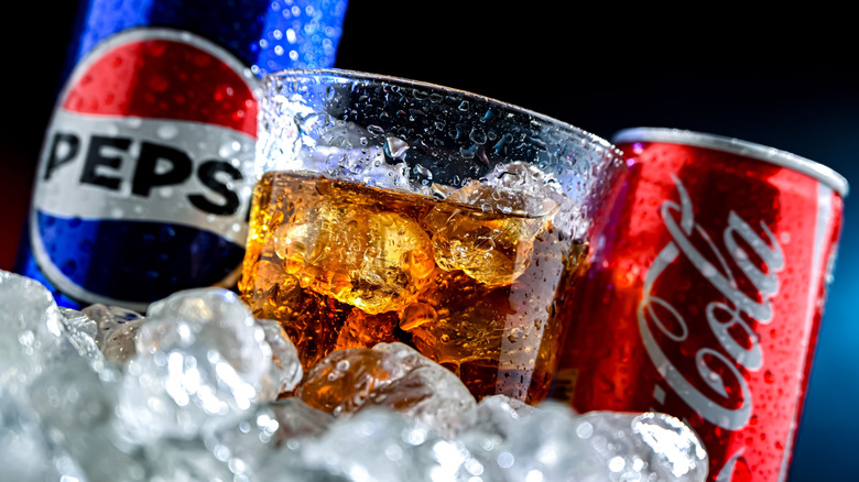 Pepsi and Coke cans with a glass of brown soda between them
