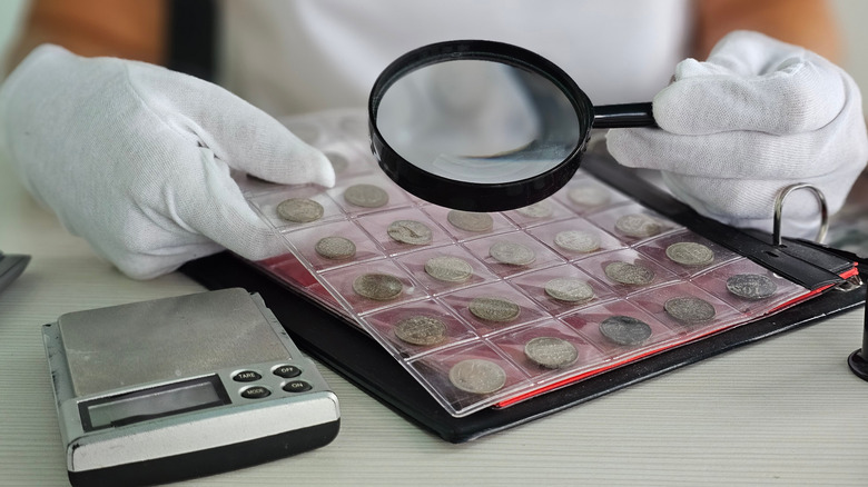 Someone with a white shirt and gloves looking at a laminated book of coins with a magnifying glass.