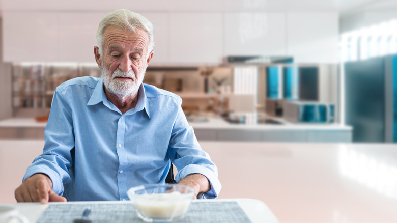 A retired man sitting alone at the kitchen table looking at a bowl of food with a sad expression.