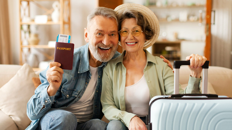 A happy senior couple seated with luggage and holding up a passport.
