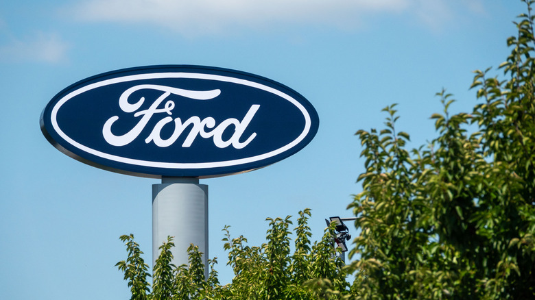 A sign displaying a Ford logo stands above a treeline on a sunny day