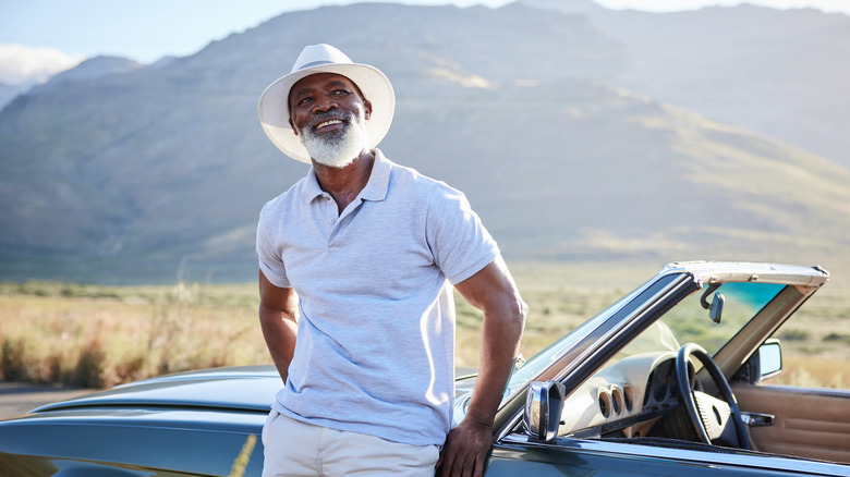 A dapper mature man leaning on his convertible and enjoying the view.