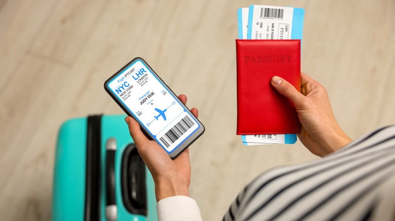 A close-up of a person's hands with one holding a phone with flight information and the other holding a passport with plane tickets inside and a blue suitcase on the floor in the background.