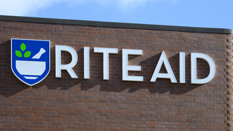A Rite Aid pharmacy store sign in the sun, on brick, against a cloudy blue sky.