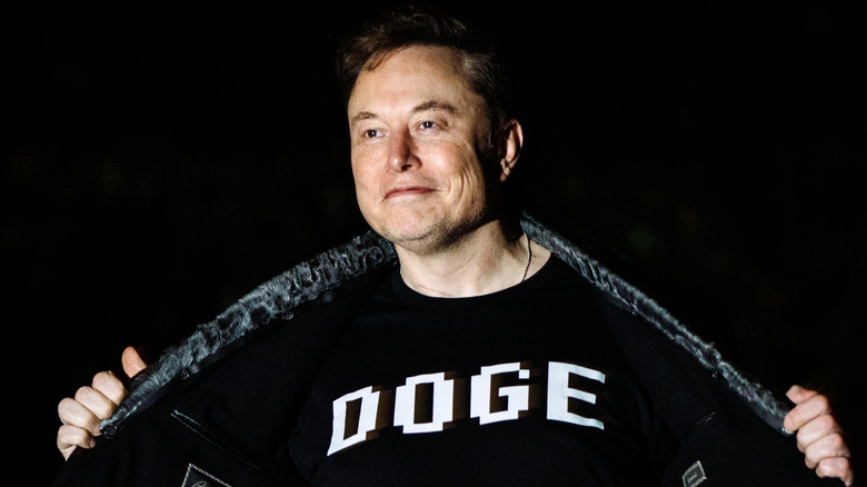 Elon Musk in bright light on a dark background, showing off a shirt that reads DOGE.