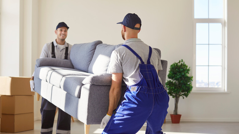Two professional movers carrying heavy gray sofa into new empty spacious room.