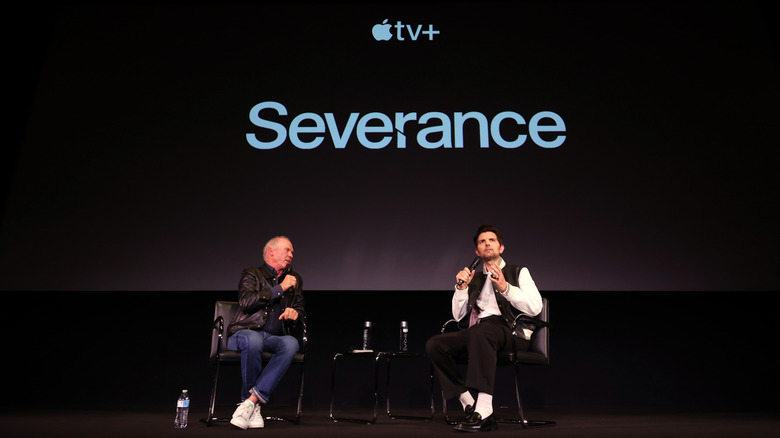 Two men sit in chairs in front of dark screen with Apple+ TV logo and word Severance on it