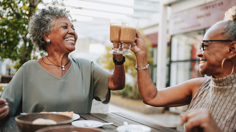 Two laughing mature women toasting a table with mugs of coffee.