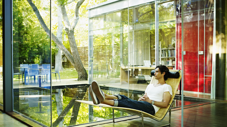 A Millennial man reclining in a chair in a bright glass walled home.