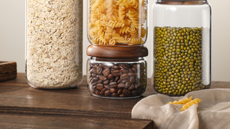 Stacked glass storage jars with acacia wood lids filled with oats pasta coffee beans and mung beans on wooden table.