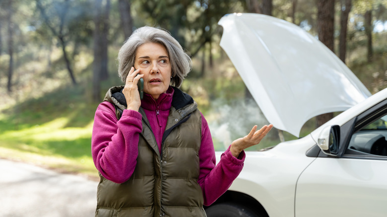 A retired woman on the phone with a white car that has the hood open in the background.