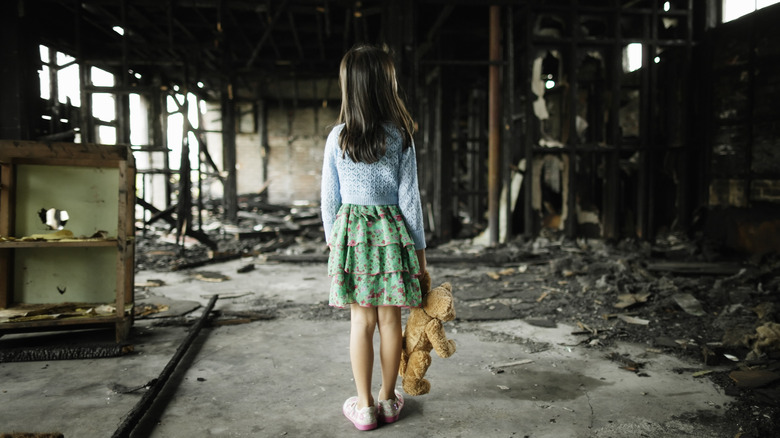 A little girl holding a teddy bear standing inside the remains of a burned home