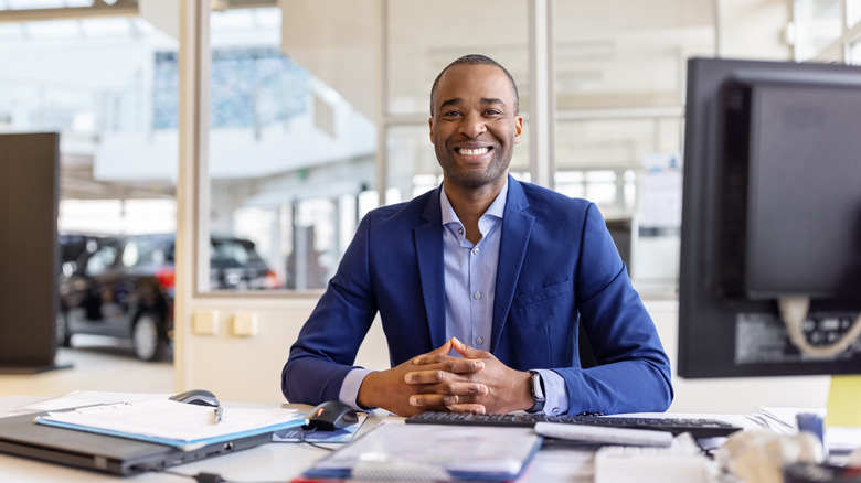 A smiling car salesman sitting behind a desk at a car dealership.