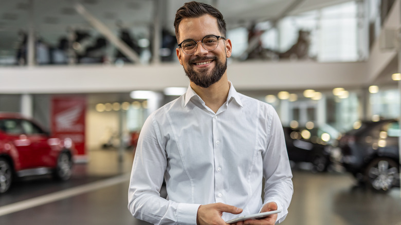 A smiling man in a crisp white shirt holding a cellphone while standing in a car dealership.