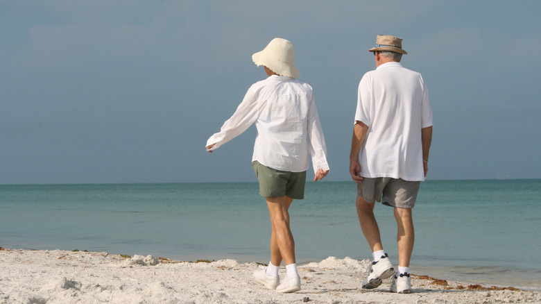 Elderly couple with white shirts walking on beach toward ocean.