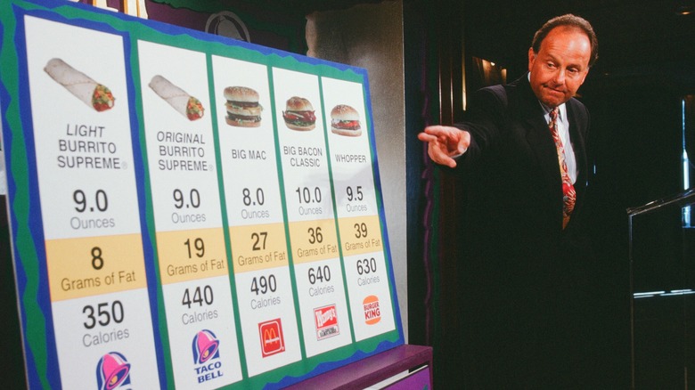 Taco Bell CEO presenting a comparison board showing Taco Bell burritos alongside the Big Mac, Big Bacon Classic, and Whopper, with listed weights, fat content, and calorie counts