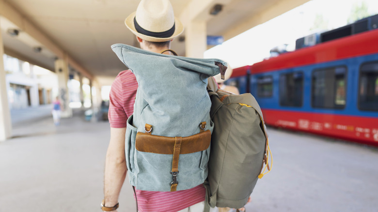 A man walking through a train station carrying several bags on his back.